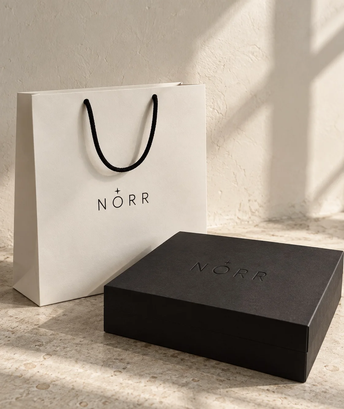 Norr packaging: bone paper bag with black rope handles and a black rigid box with the debossed wordmark, in soft window light (AI mockup)