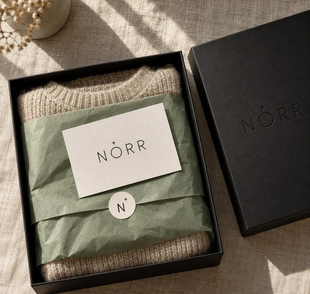 Open black Norr box: an oat knit wrapped in sage tissue, a card and a star seal (AI mockup)